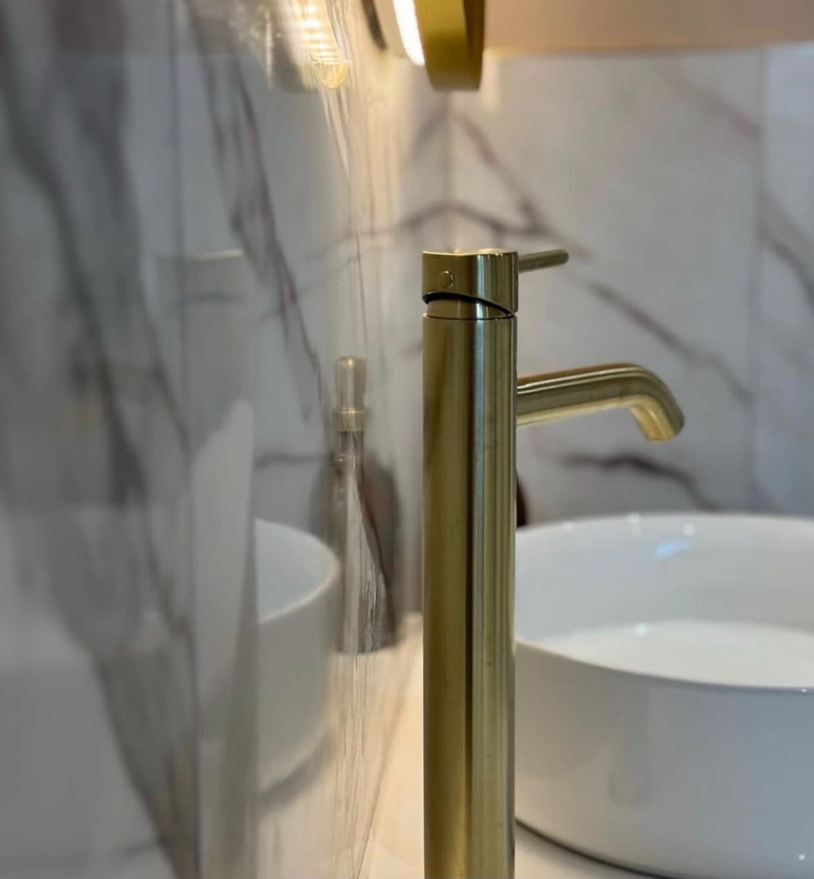 Close-up of a running brushed brass tall monobloc tap beside a round white vessel sink with marble-tiled splashback