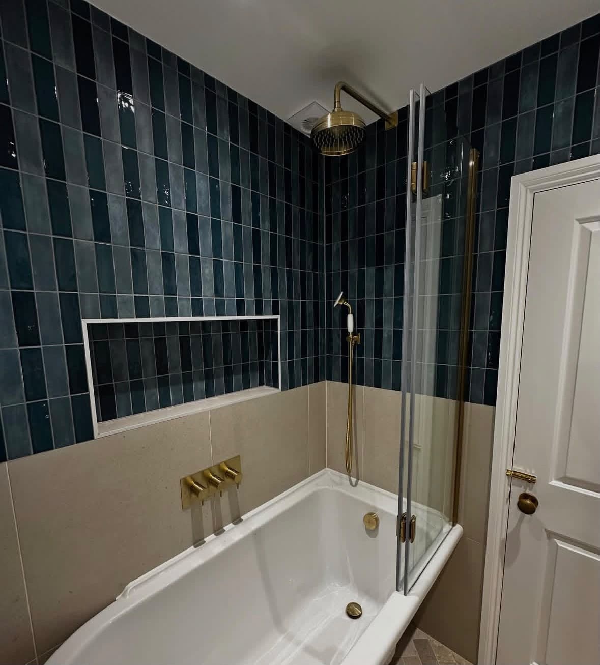 Bathroom with deep teal vertical brick tiles, bath with folding glass screen, brass overhead rain shower and brass mixer taps